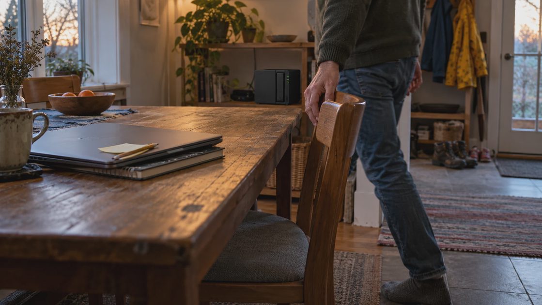 Person pushing a dining chair toward a table with a closed laptop and notebook, with shoes by the door.